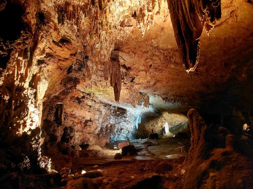 A dimly lit cave interior with stalactites hanging from the ceiling and rocky formations on the ground.