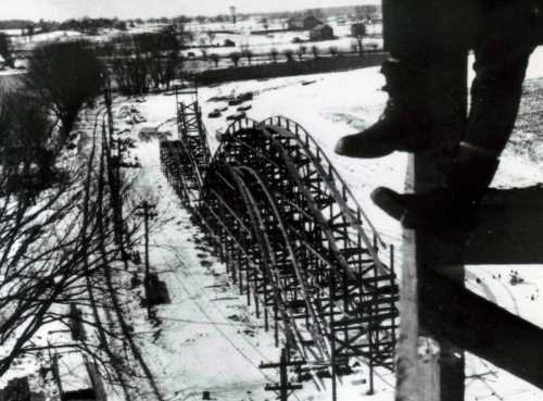 A black-and-white photo of a snowy landscape with a partially constructed roller coaster and a person's boot in the foreground.