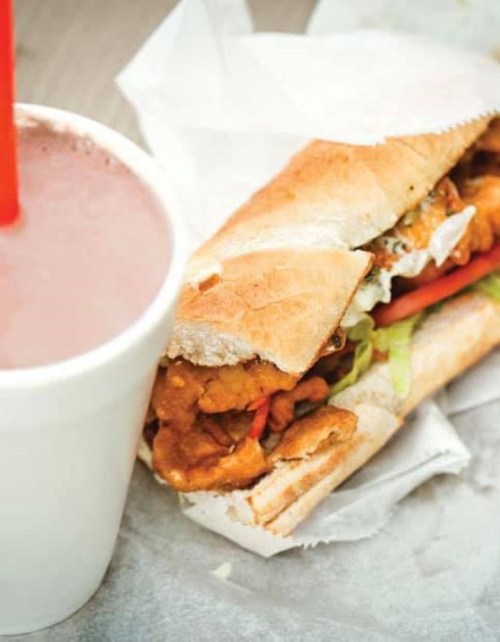 A close-up of a sandwich filled with fried chicken, lettuce, and peppers, alongside a cup of pink beverage.