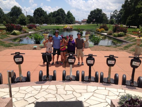 A group of five people poses with segways in front of a park with a pond and colorful flowers on a sunny day.