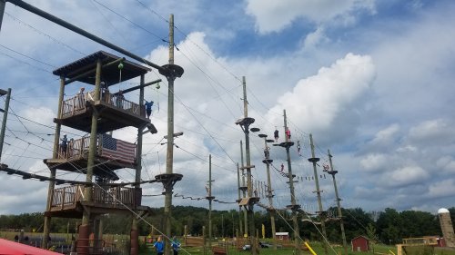 A high ropes course with wooden platforms and cables, set against a cloudy sky and green landscape.
