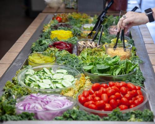 A vibrant salad bar with various fresh vegetables, including cucumbers, tomatoes, and greens, with tongs for serving.