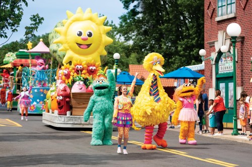 A colorful parade featuring cheerful characters, including Big Bird and other beloved puppets, along a festive street.