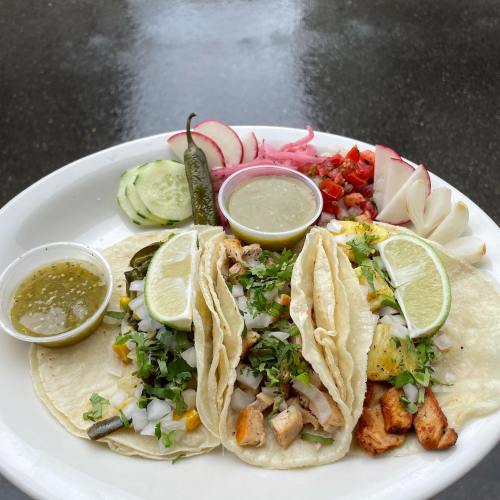 A plate of three tacos topped with chicken, onions, and cilantro, served with lime, salsa, and fresh vegetables.
