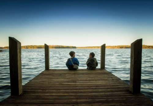 Two children sit on a wooden dock, gazing at a calm lake under a clear blue sky.