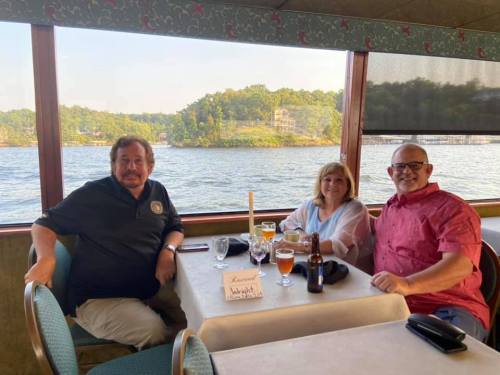Three people sit at a table by a window, enjoying drinks with a scenic view of water and hills in the background.