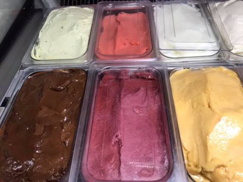 A variety of gelato flavors in clear containers, including green, pink, brown, purple, and yellow.