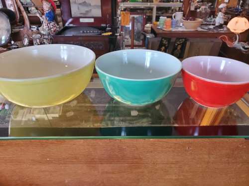 Three colorful mixing bowls in yellow, green, and red, displayed on a glass shelf in a vintage shop.