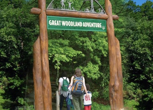 A family approaches a wooden archway sign that reads "Great Woodland Adventure," surrounded by lush greenery.