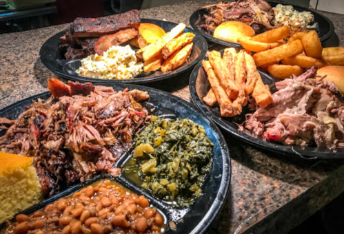 A spread of barbecue dishes including pulled pork, sides of greens, baked beans, cornbread, and various fried items.