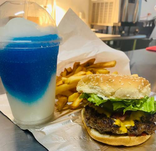 A sesame seed burger with lettuce and cheese, served with fries and a blue slushy drink on a silver surface.