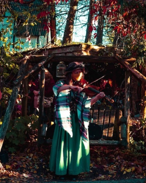 A woman in a green dress plays the violin outdoors, surrounded by autumn leaves and a rustic wooden structure.