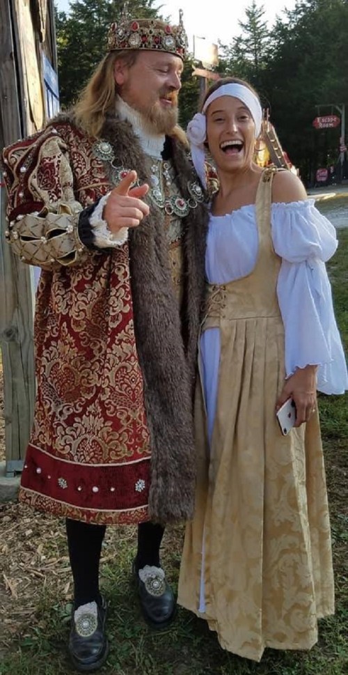 A man in a royal costume and a woman in a medieval dress smile together outdoors.