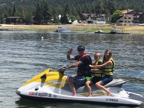 A man and two children wave while riding a yellow jet ski on a lake, surrounded by trees and houses in the background.