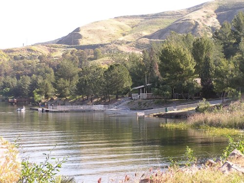 A serene lakeside view with trees, a dock, and gentle hills in the background under a clear sky.