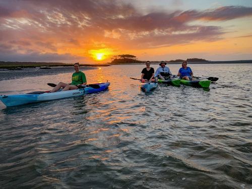 Four people kayaking at sunset, with vibrant colors reflecting on the water and a serene landscape in the background.