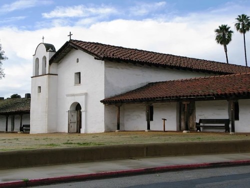 Historic white church with a red-tiled roof, surrounded by palm trees and a clear blue sky.