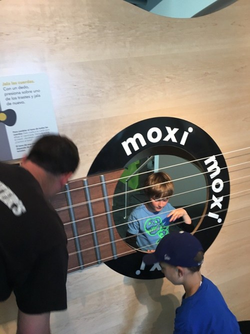Two children interact with a large guitar display at a museum, while an adult leans in to assist.