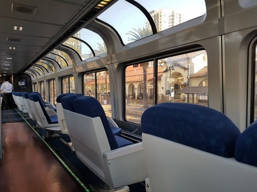 Interior of a train with blue seats and large windows, showing a view of a station and palm trees outside.
