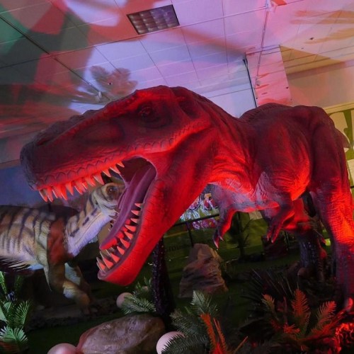 A large, red dinosaur model with an open mouth, surrounded by greenery and eggs in a brightly lit exhibit.