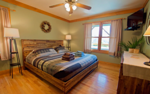 Cozy bedroom with a wooden bed, plaid bedding, lamps, and a window with a view, featuring warm tones and natural decor.