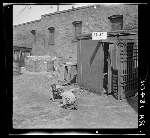 Two children playing on the ground near an outdoor toilet in a brick building setting.