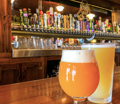 Two glasses of beer sit on a wooden bar, with a colorful array of tap handles in the background.