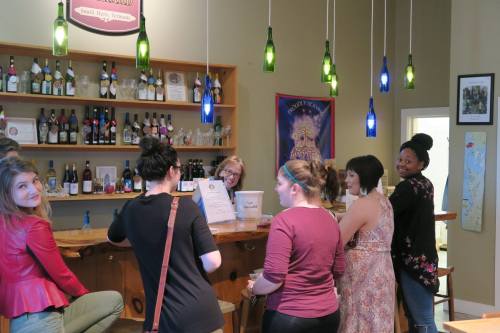 A group of women at a bar, chatting and enjoying drinks, with colorful pendant lights and a wine display in the background.
