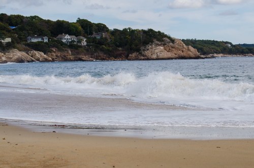 Waves gently crash on a sandy beach with rocky cliffs and houses in the background under a cloudy sky.