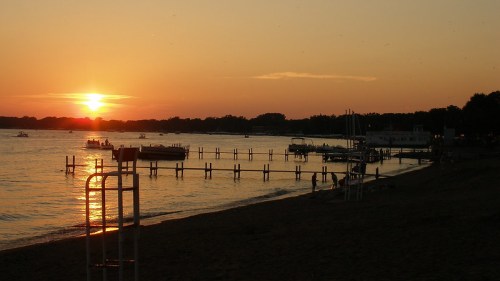 Sunset over a calm lake, with boats on the water and a sandy beach in the foreground. Silhouettes of trees in the distance.