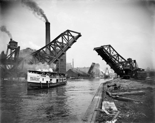 A historic black-and-white photo of a steam boat navigating under raised drawbridges on a river.