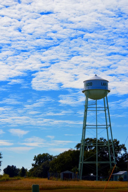 A green water tower labeled "Placid Lake" stands against a blue sky with scattered clouds and a grassy field below.