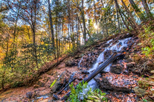 A serene waterfall cascades over rocks, surrounded by autumn foliage and sunlight filtering through trees.