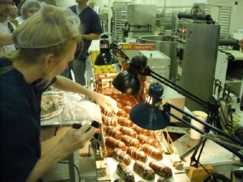 A person in a hairnet drizzles chocolate over pastries in a factory, with bright lights illuminating the workspace.