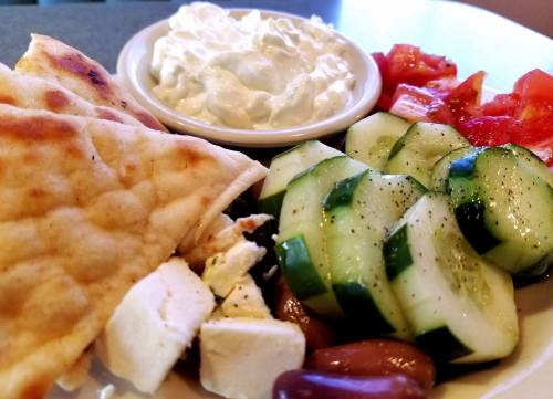A plate with pita bread, cucumbers, tomatoes, olives, feta cheese, and a bowl of creamy dip.