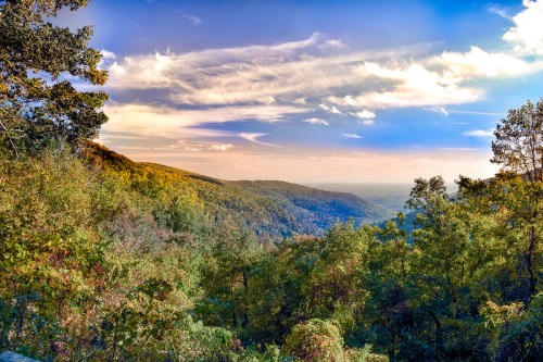 A scenic view of rolling hills and valleys under a colorful sky, surrounded by lush green trees.
