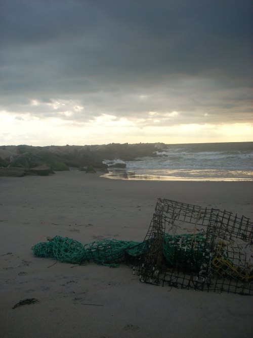 A beach scene with dark clouds, gentle waves, and a fishing trap lying on the sand near rocky formations.
