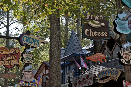Colorful food signs, including "Fried Cheese" and "Crepes," amidst trees and charming buildings at a festival.