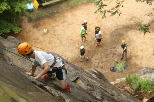 A climber ascends a rock wall while others observe from below, wearing helmets and safety gear in a forested area.