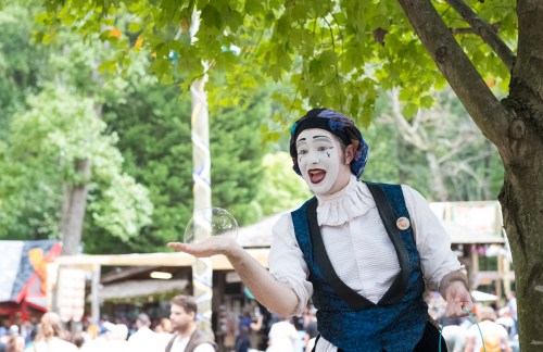 A mime in a blue costume performs under a tree, playfully holding a bubble in one hand at a lively outdoor festival.