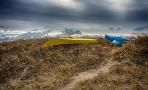 A yellow canoe and a blue kayak resting on a sandy dune with tall grass under a cloudy sky.