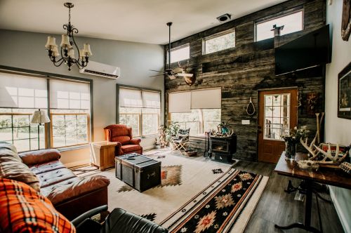 Cozy living room with large windows, wooden walls, a fireplace, and comfortable seating. Rustic decor and a warm atmosphere.