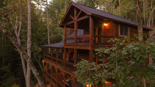 A rustic wooden cabin nestled among tall trees, featuring a spacious deck and warm lighting at dusk.