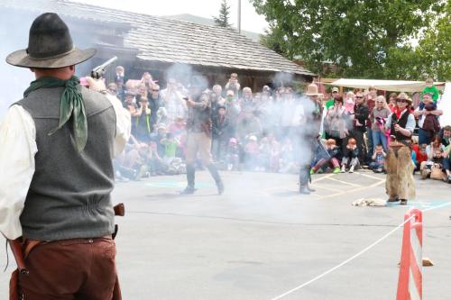 A cowboy show with performers in period costumes, gunfire, and a crowd watching in a festive outdoor setting.