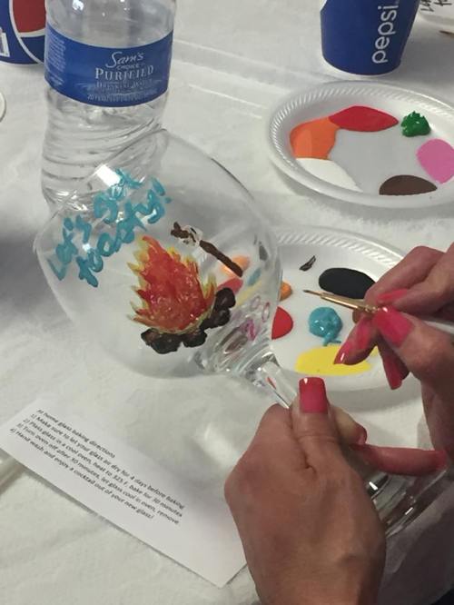 A person paints a colorful design on a wine glass, with paint palettes and a water bottle in the background.