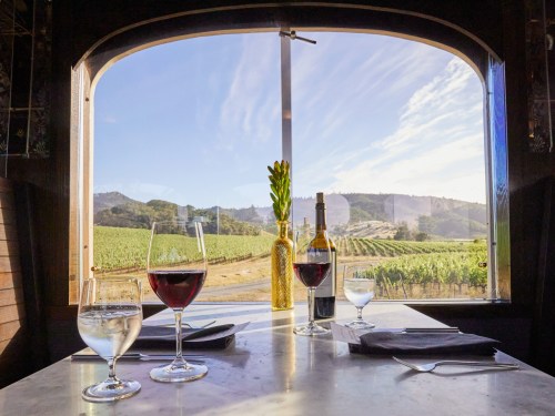 A scenic view from a restaurant window overlooking vineyards, with glasses of red and white wine on the table.