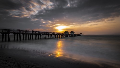 A serene sunset over a pier, with soft waves and clouds creating a tranquil atmosphere.