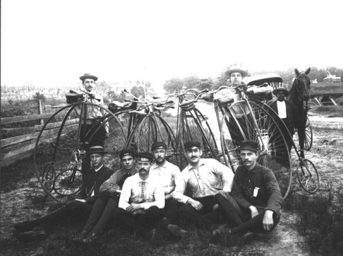 A group of men poses with vintage bicycles and a horse, set in a rural landscape.