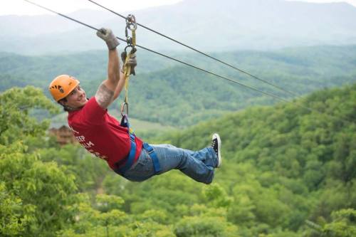 A person in a red shirt and helmet zip-lining over a lush green landscape with mountains in the background.