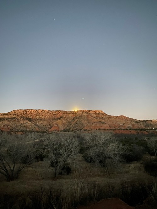 A serene landscape at dusk, featuring a mountain with a glowing peak and bare trees in the foreground.